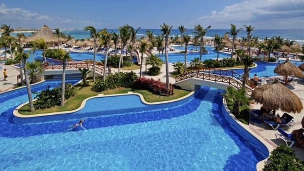 Zona de piscina en un complejo turístico con varias piscinas, palmeras y vista a la playa.