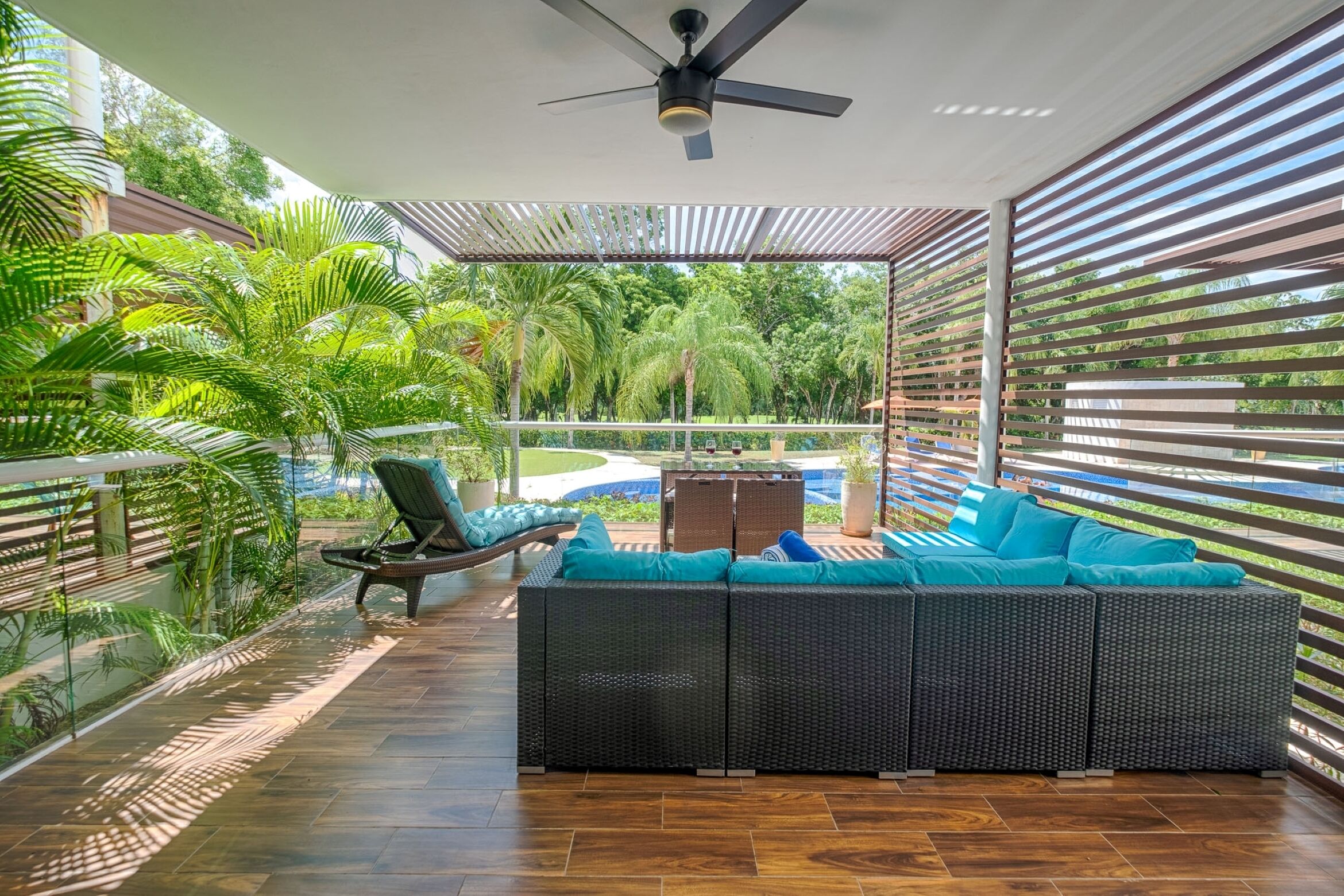 An outdoor seating area with a sofa and chairs, a ceiling fan, and a view of a pool and greenery.