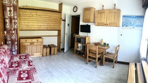 A compact living space with a wooden sofa, a dining table with chairs, a small kitchen area, and a mounted TV. - Studio Dans une Petite Copropriété en Plein Centre de Tignes (Tignes)