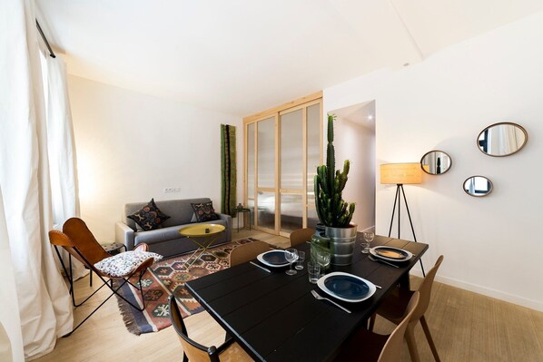 A modern living room with a dining area, a grey sofa with patterned cushions, a cactus plant, and a table set for two.