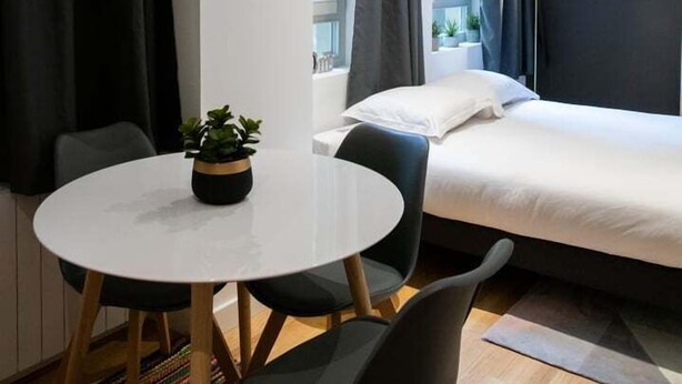 Una habitación de hotel moderna con una cama, una mesa redonda con sillas, una planta en maceta y un ventanal con cortinas.