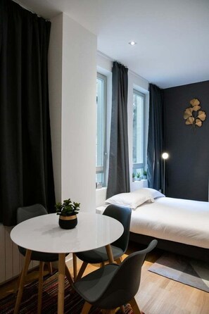 Une chambre d’hôtel moderne, avec un lit, une table ronde entourée de chaises, une plante en pot et une fenêtre avec des rideaux.