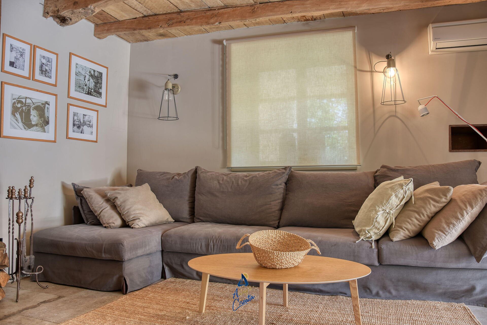 A cozy living room with a grey sofa, a round wooden coffee table, and a woven basket. There are framed pictures on the wall, a hanging light fixture, and a window with blinds.