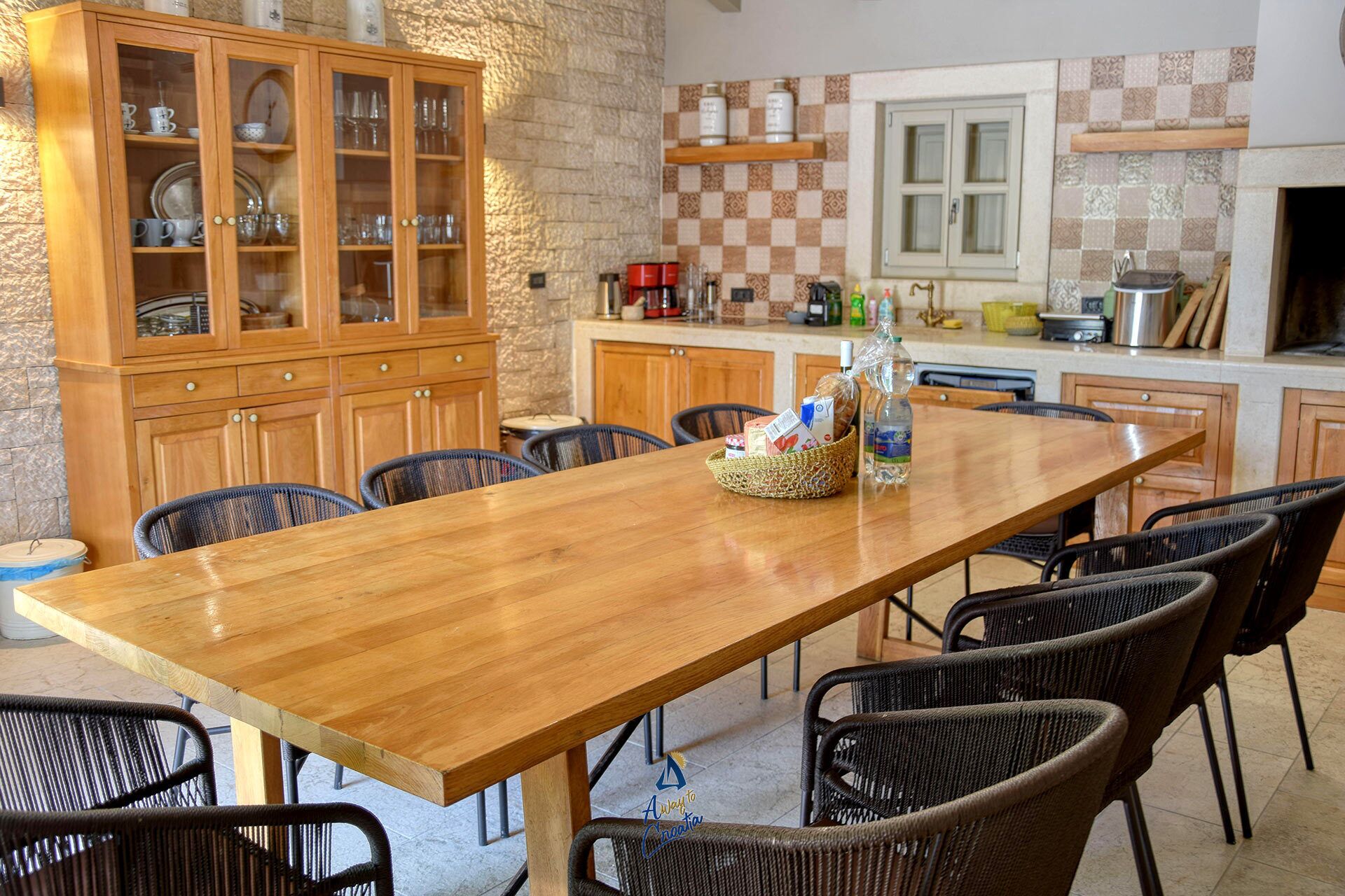 A wooden dining table with wicker chairs, a stone wall, and a kitchen with checkered tiles and wooden cabinets.