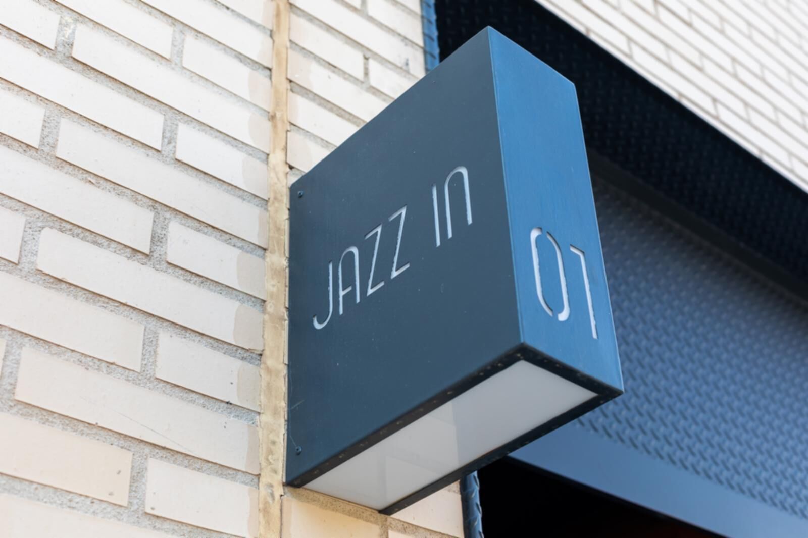 Segno nero con scritta bianca che riporta "JAZZ IN 01" fissato a un muro in mattoni.