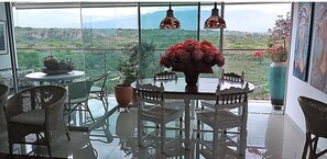 A dining area with a table and chairs, a vase with red flowers, and a view of the landscape through large windows. - Casa Chic! (Neiva)