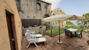 A patio with a table and chairs, a large umbrella, and a rustic building in the background. - Tangerine house by the sea (Riposto)