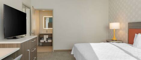 A hotel room with a bed, a TV, a desk, a lamp, and a bathroom visible through an open door.