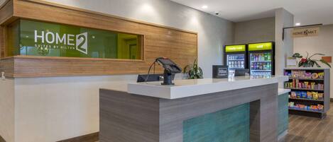 A reception desk with a sign reading "HOME 2 SUITES BY HILTON" and a digital screen. There are two beverage dispensers labeled "fity cold" and "cold." A plant and a shelf with snacks are visible in the background.
