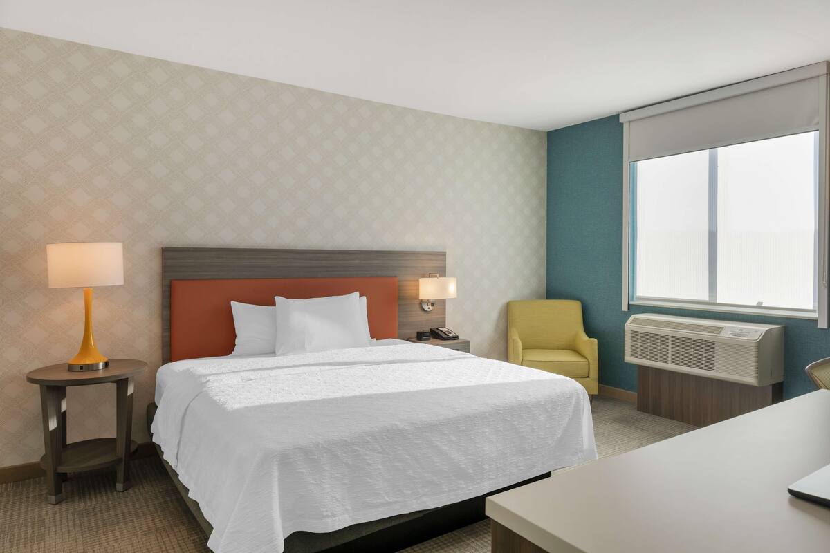 Una habitación de hotel con cama, mesita de noche con lámpara, sillón, aire acondicionado y ventana con persianas.