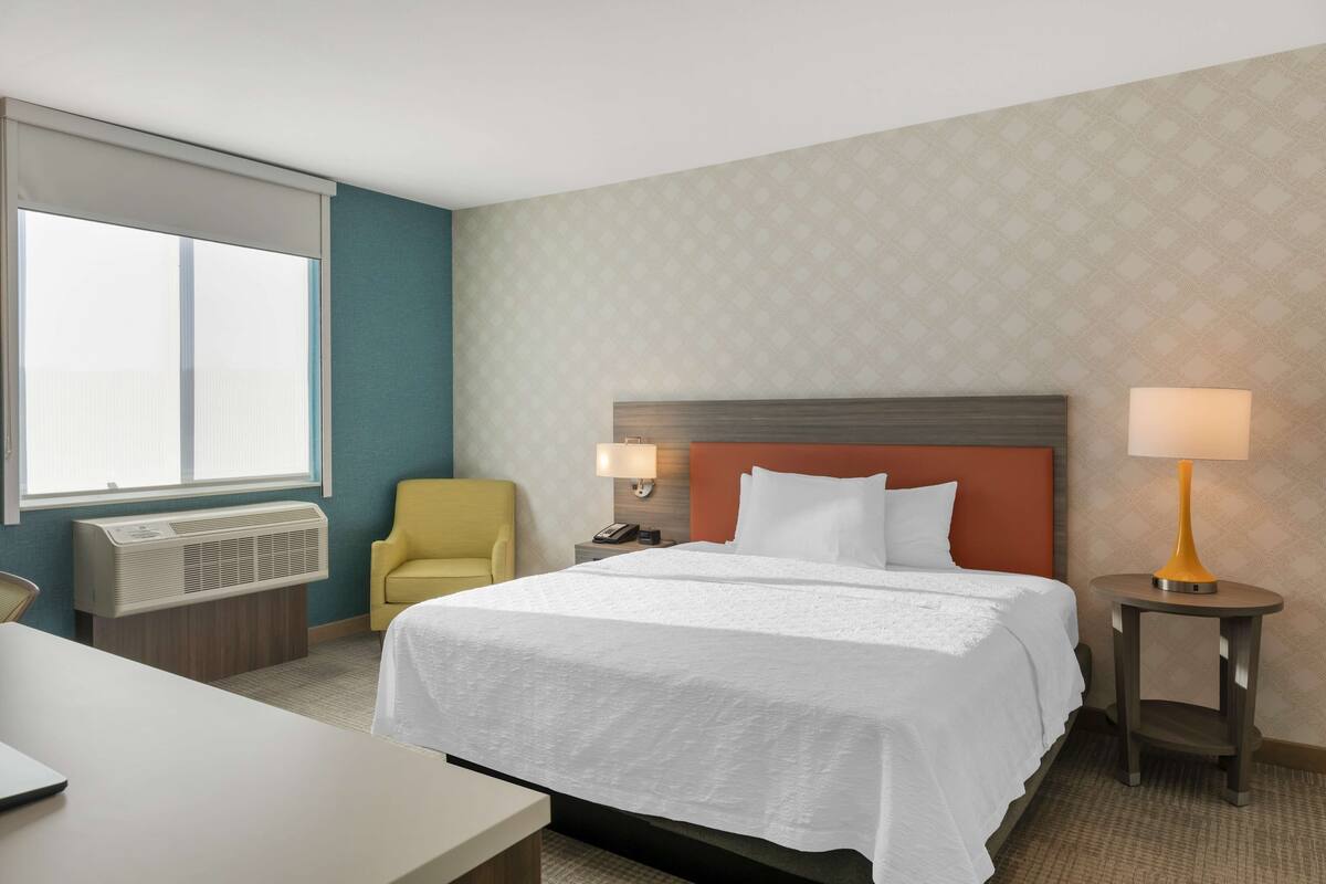 Habitación de hotel con cama, escritorio, silla, mesita de noche con lámpara y ventana con persianas.