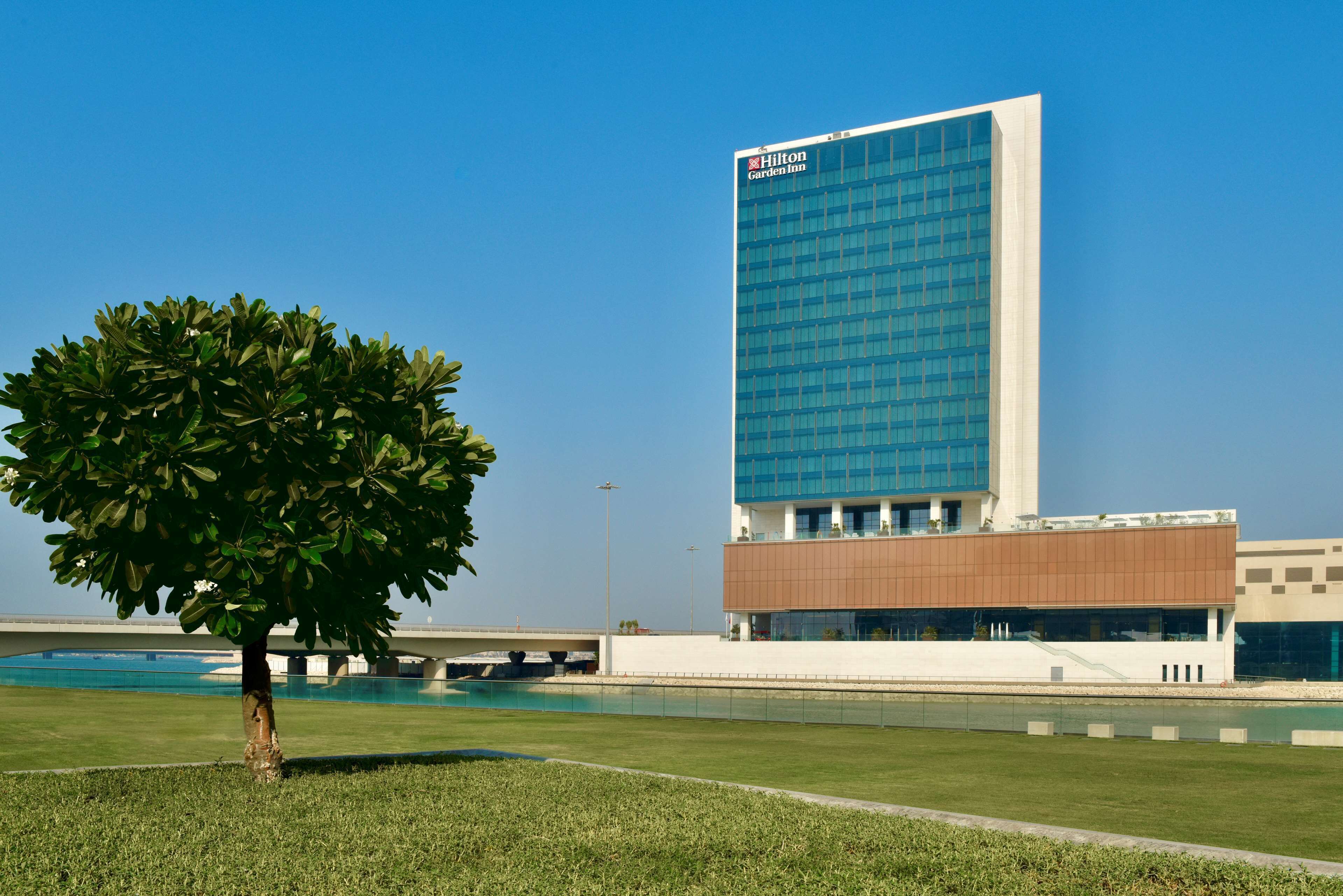 Un bâtiment moderne, sur lequel on peut voir l’enseigne d’un hôtel Hilton Garden Inn, un grand arbre au premier plan et un ciel bleu dégagé.