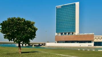 Un bâtiment moderne, sur lequel on peut voir l’enseigne d’un hôtel Hilton Garden Inn, un grand arbre au premier plan et un ciel bleu dégagé.