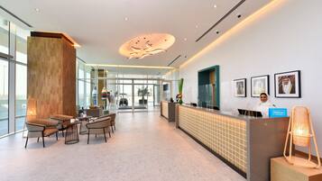 A modern hotel reception area with a wooden reception desk, seating area with chairs, and a large window offering a view of the outside.