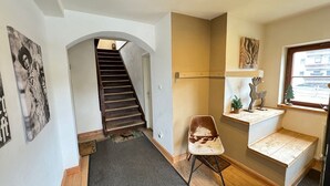 Uma escadaria leva a um andar superior, com uma cadeira, um pequeno armário com objetos decorativos e uma janela com vista.