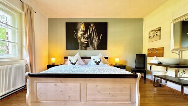 Quarto com uma cama grande, abajures, um quadro na parede, piso de madeira e uma janela com cortinas.