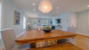 A spacious dining area with a wooden table, striped chairs, a chandelier, and a view of a kitchen and living area. - Blue Haven Home, Fire Pit & Blocks from Beach (Cape Charles)