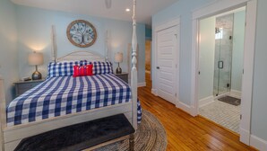 A bedroom with a checkered bedspread, a round wall clock, a bedside lamp, and a bench. - Blue Haven Home, Fire Pit & Blocks from Beach (Cape Charles)