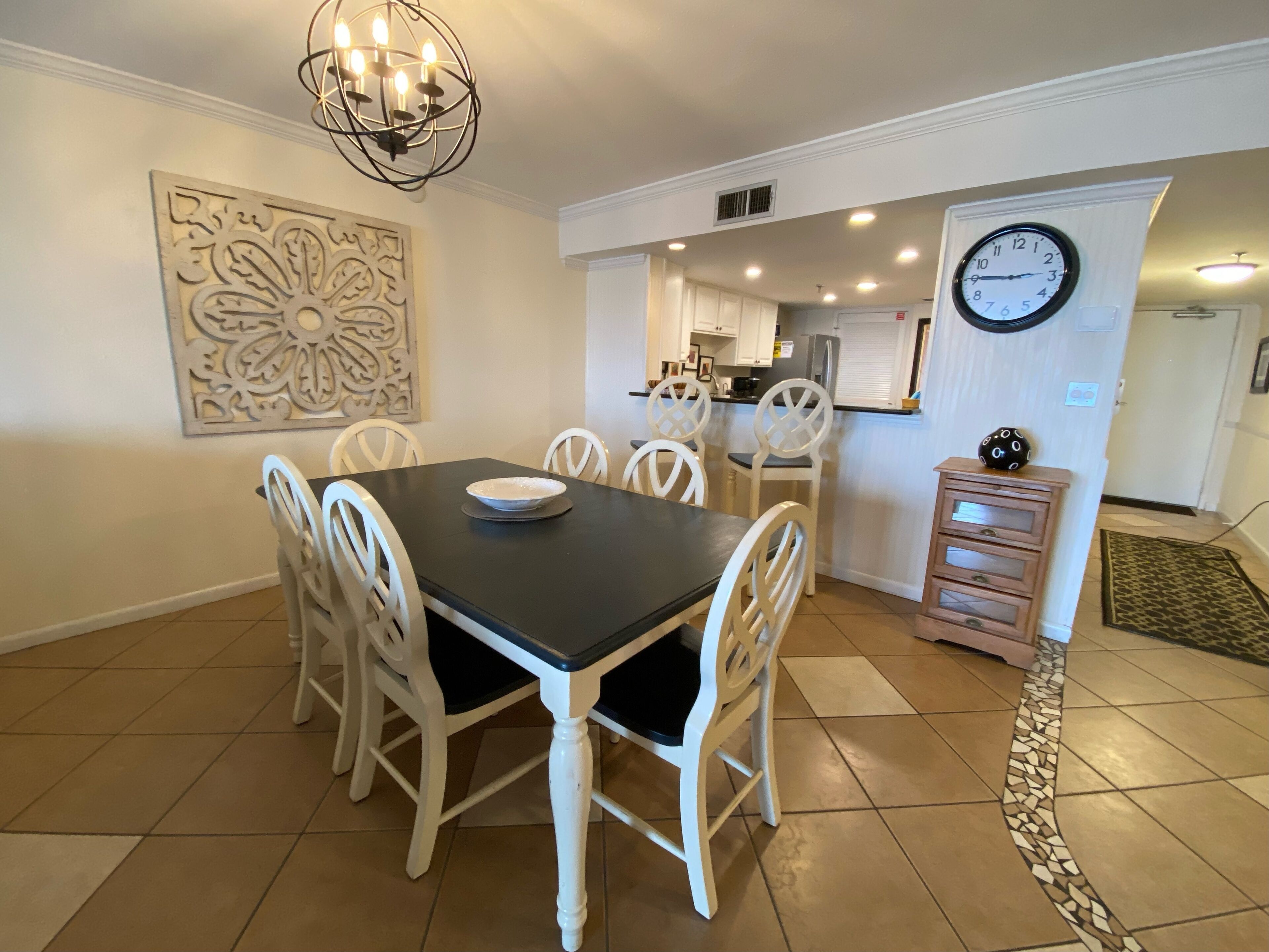 A dining area with a black table and white chairs, a decorative wall piece, and a clock on the wall.