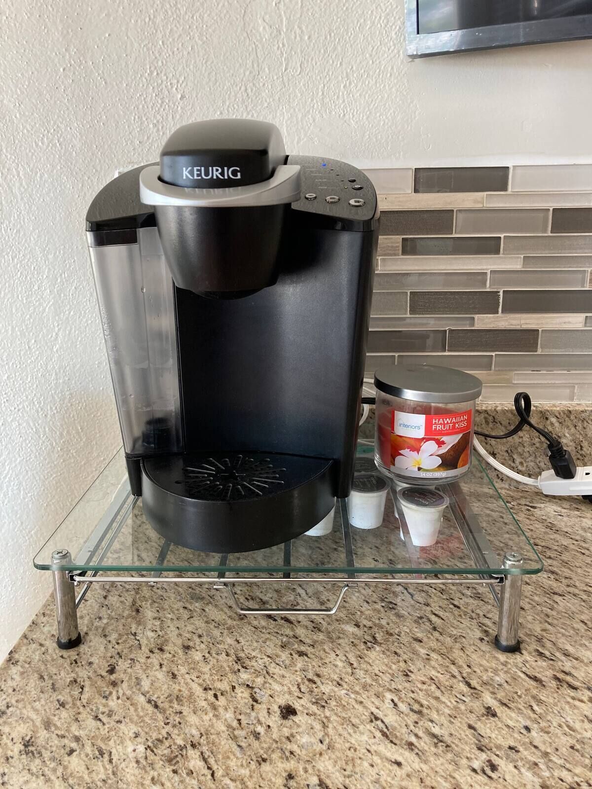 A Keurig coffee maker on a glass shelf with a container of Hawaiian Fruit Kiss coffee pods.