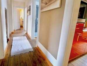 A hallway with wooden flooring, white walls, and a red area that appears to be a kitchen or dining space.