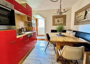 A kitchen with red cabinets, a wooden dining table, and a bench. There is a chandelier, a painting on the wall, and a plant on the table. - Kitz Apartment 3 mit Garten, Nähe Schwarzsee (Kitzbühel)