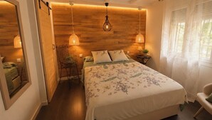 Een slaapkamer met een houten hoofdeinde, een bed met bloemenbeddels, twee bedlampen, een kleine tafel met een plant en een stoel.