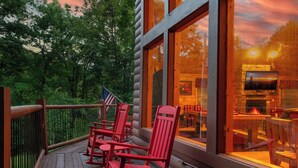 Een houten terras met rode Adirondack-stoelen, een uitzicht op het interieur van een blokhut met een open haard en een Amerikaanse vlag.