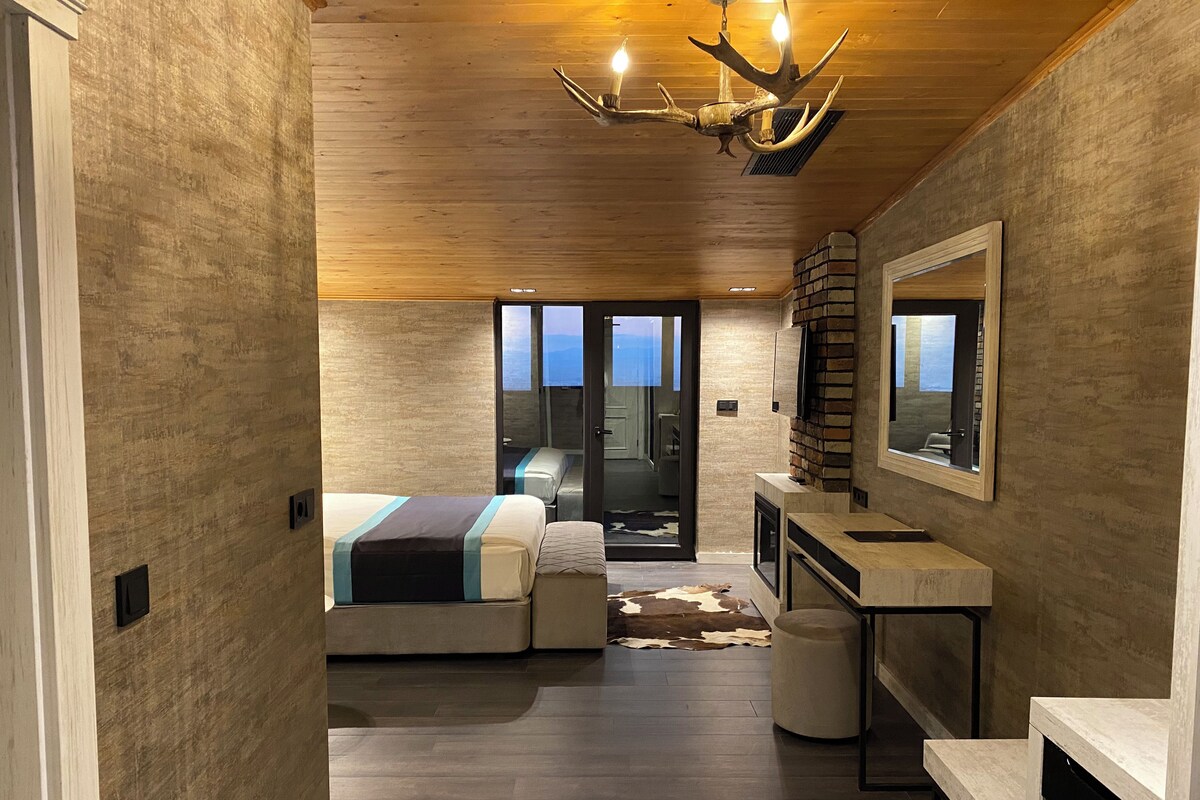 A modern hotel room with a wooden ceiling, a bed with a headboard, a small seating area, a television, and a fireplace.