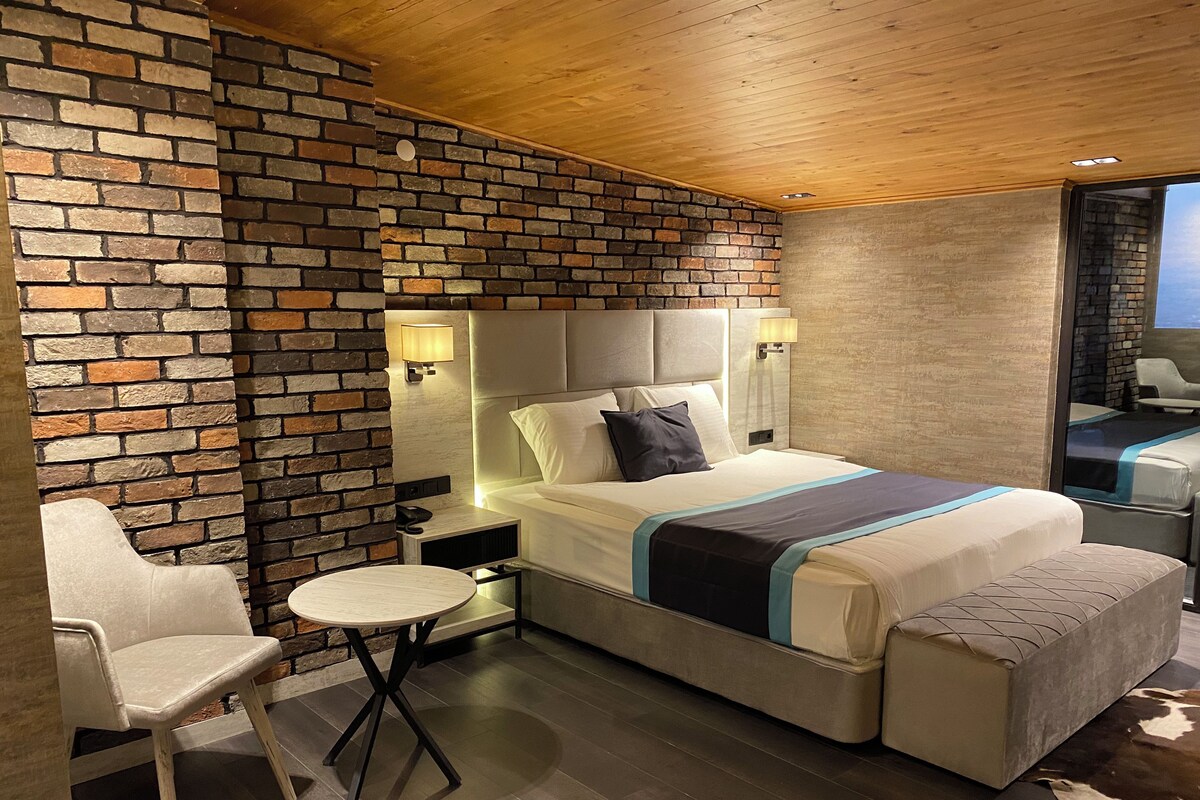 A modern hotel room with a brick accent wall, a large bed with a headboard, bedside tables with lamps, a small round table, and a chair.