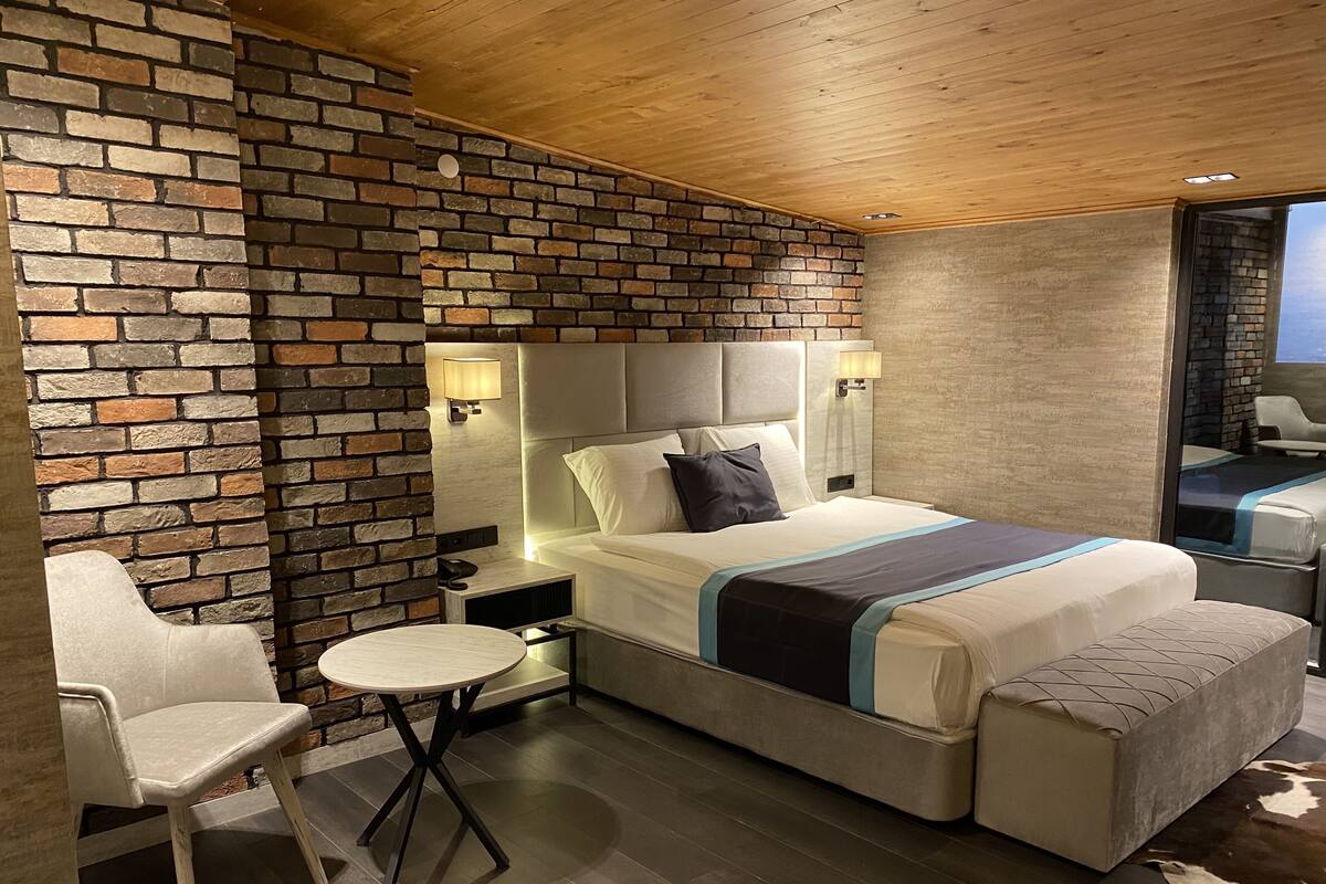 A modern hotel room with a brick accent wall, a large bed with a headboard, bedside tables with lamps, a small round table, and a chair.
