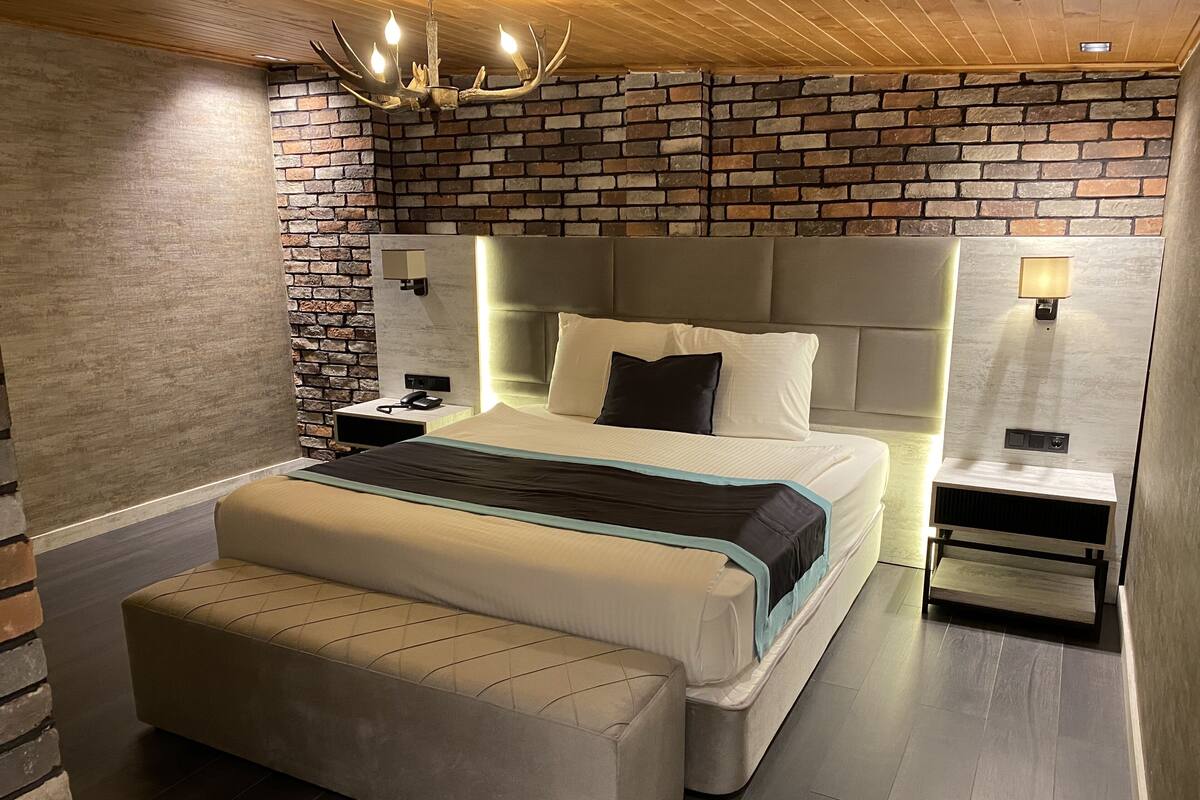 A modern bedroom with a brick accent wall, a large bed with a headboard, bedside lamps, and a wooden floor.