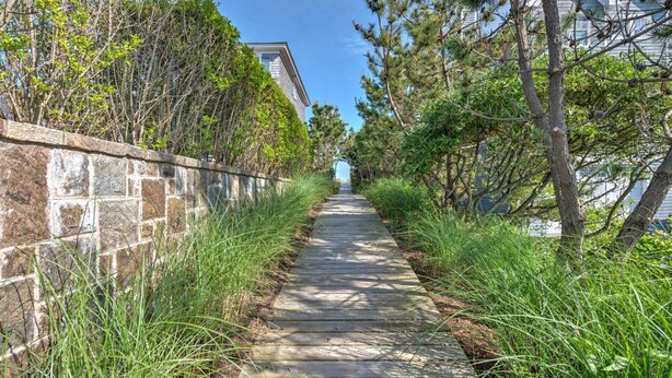A wooden pathway flanked by stone walls and dense greenery, leading towards a building with a visible window. - Bungalow 2 - 1br, Water View (Westhampton Beach)