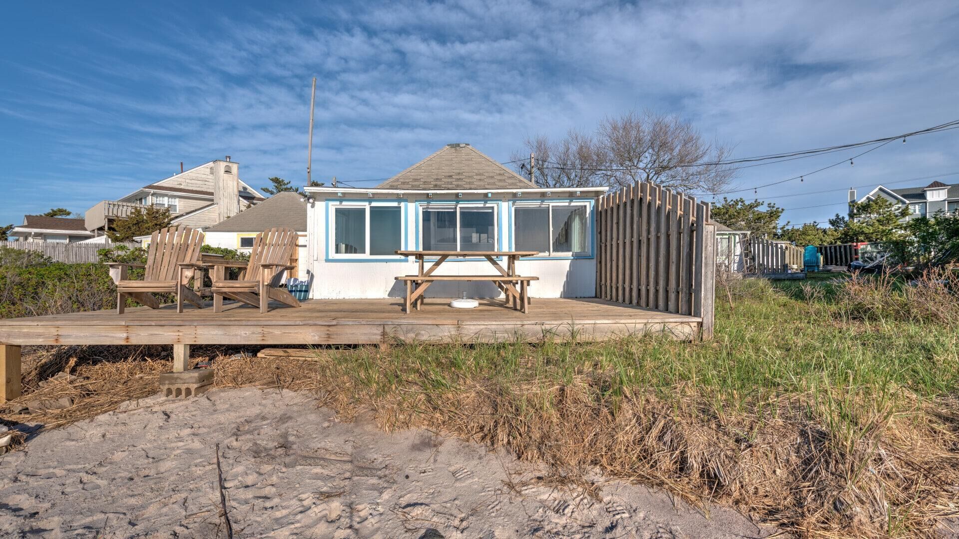 A beach house with a wooden deck, a picnic table, and two Adirondack chairs.