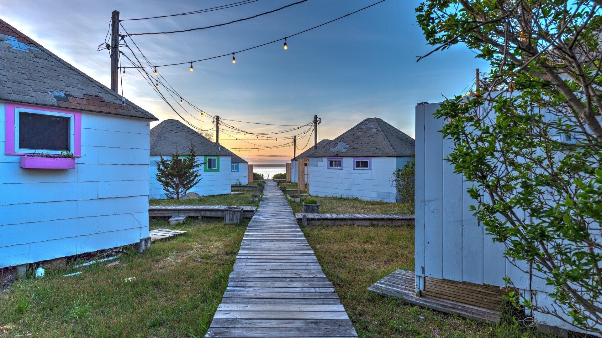 A wooden pathway leading to a row of small, single-story cabins with a clear sky above.