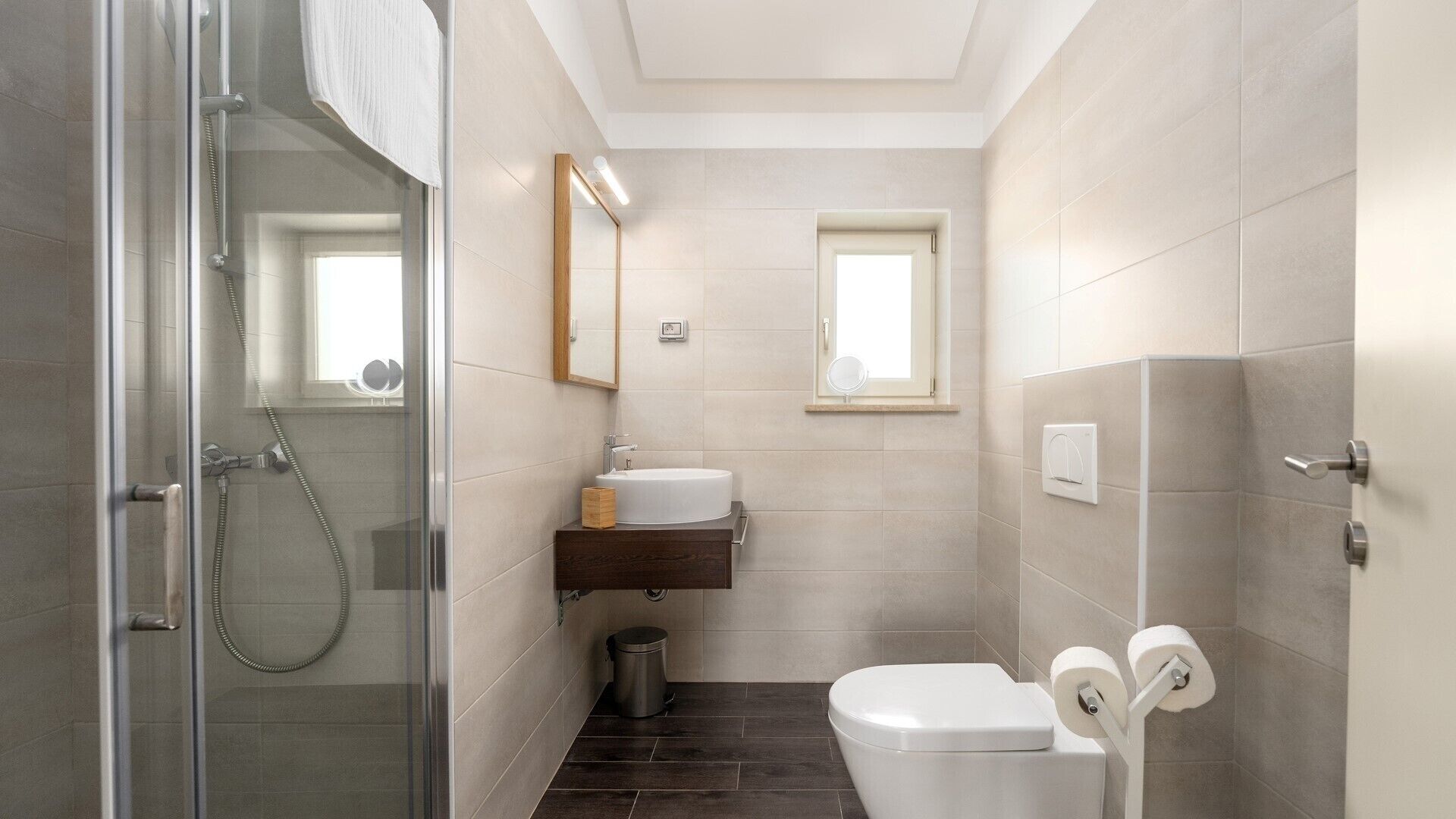 A compact bathroom with a glass shower enclosure, a sink with a mirror above, a toilet with toilet paper, and a small window.