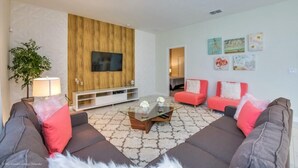 A modern living room with a flat-screen TV mounted on a wooden panel wall, a glass coffee table, two grey sofas with red cushions, and a red sofa with a white throw. - 5 Bed Pool With Overflowing Spa In Champions Gate! 5 Bedroom Villa by RedAwning (Davenport)