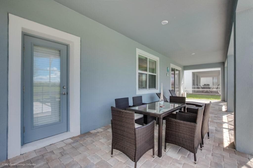 A covered patio area with a glass-top table and wicker chairs, a door with a window, and a view of a lawn and building in the background.