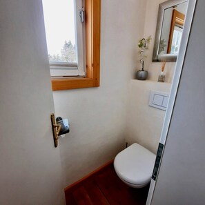 Een kleine badkamer met toilet, raam, spiegel en een lichtschakelaar op de muur.