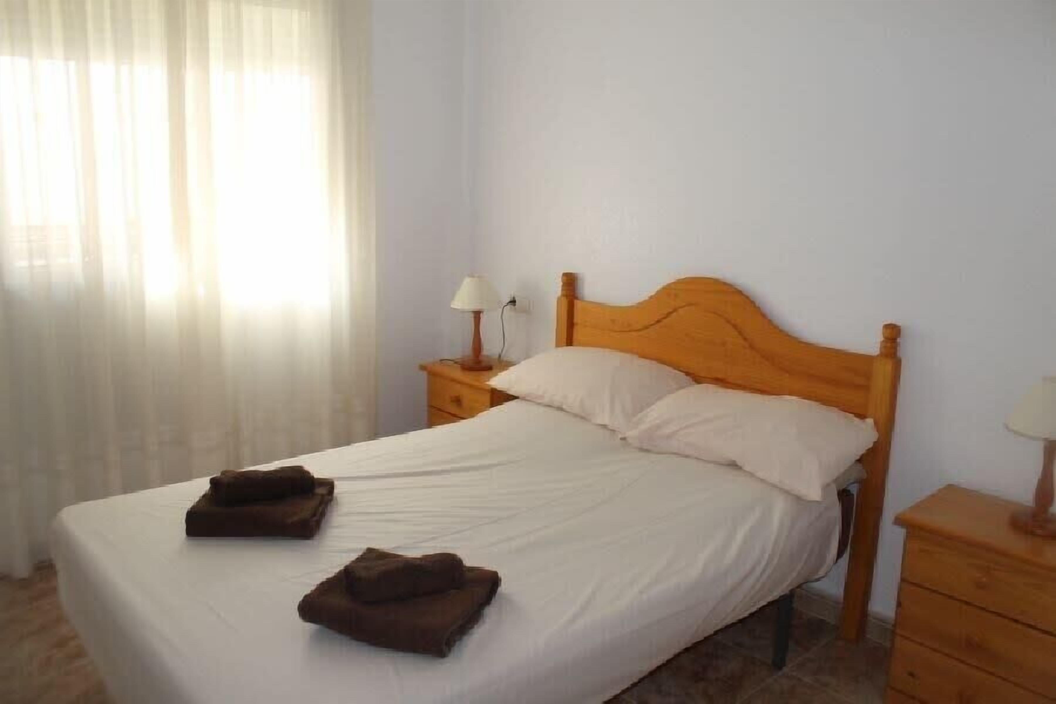 A neatly made bed with white linens and two folded brown towels, a wooden headboard, and a bedside table with a lamp.