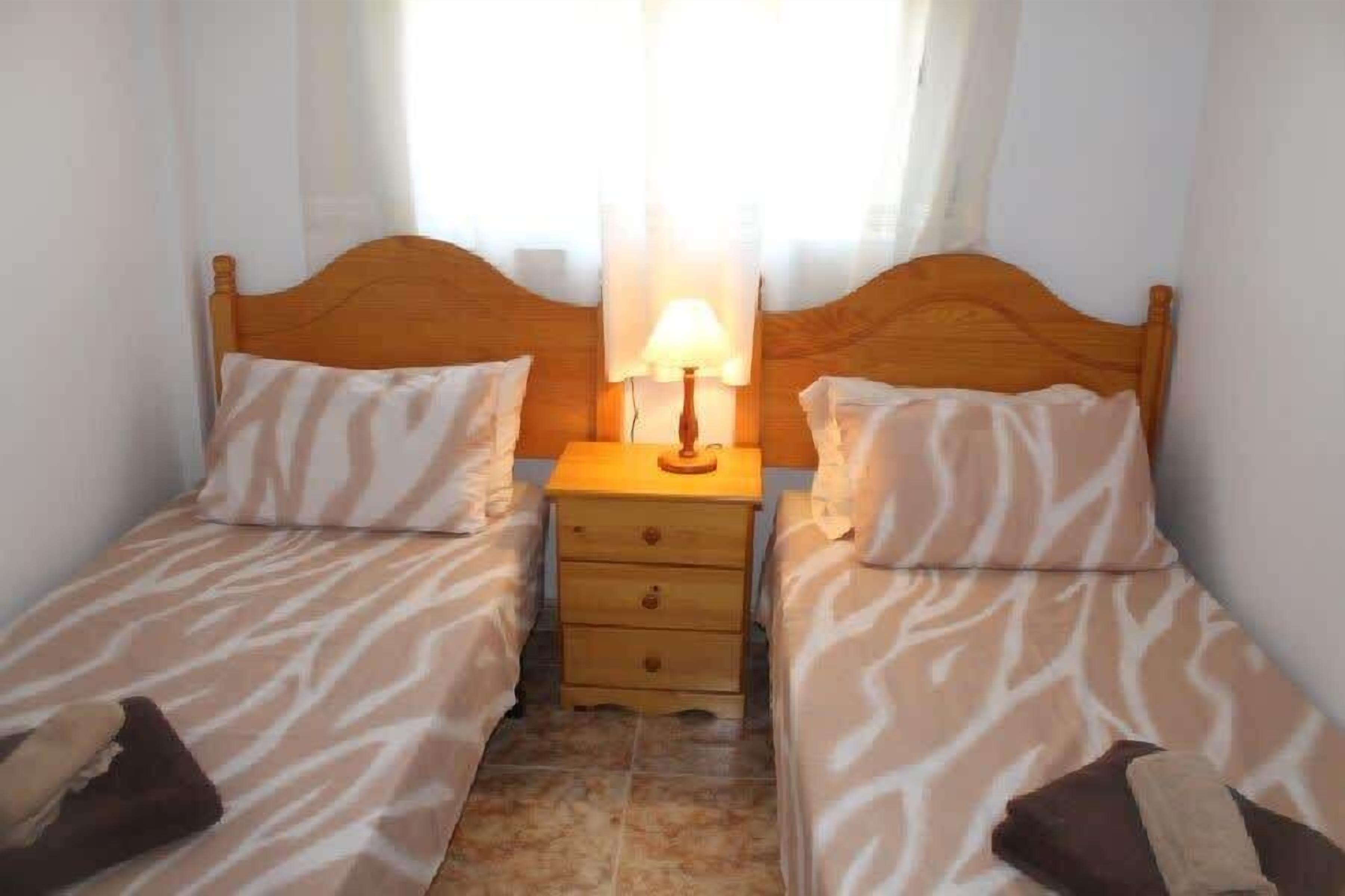 A small, clean room with two single beds, a nightstand with a lamp, and a window with sheer curtains.
