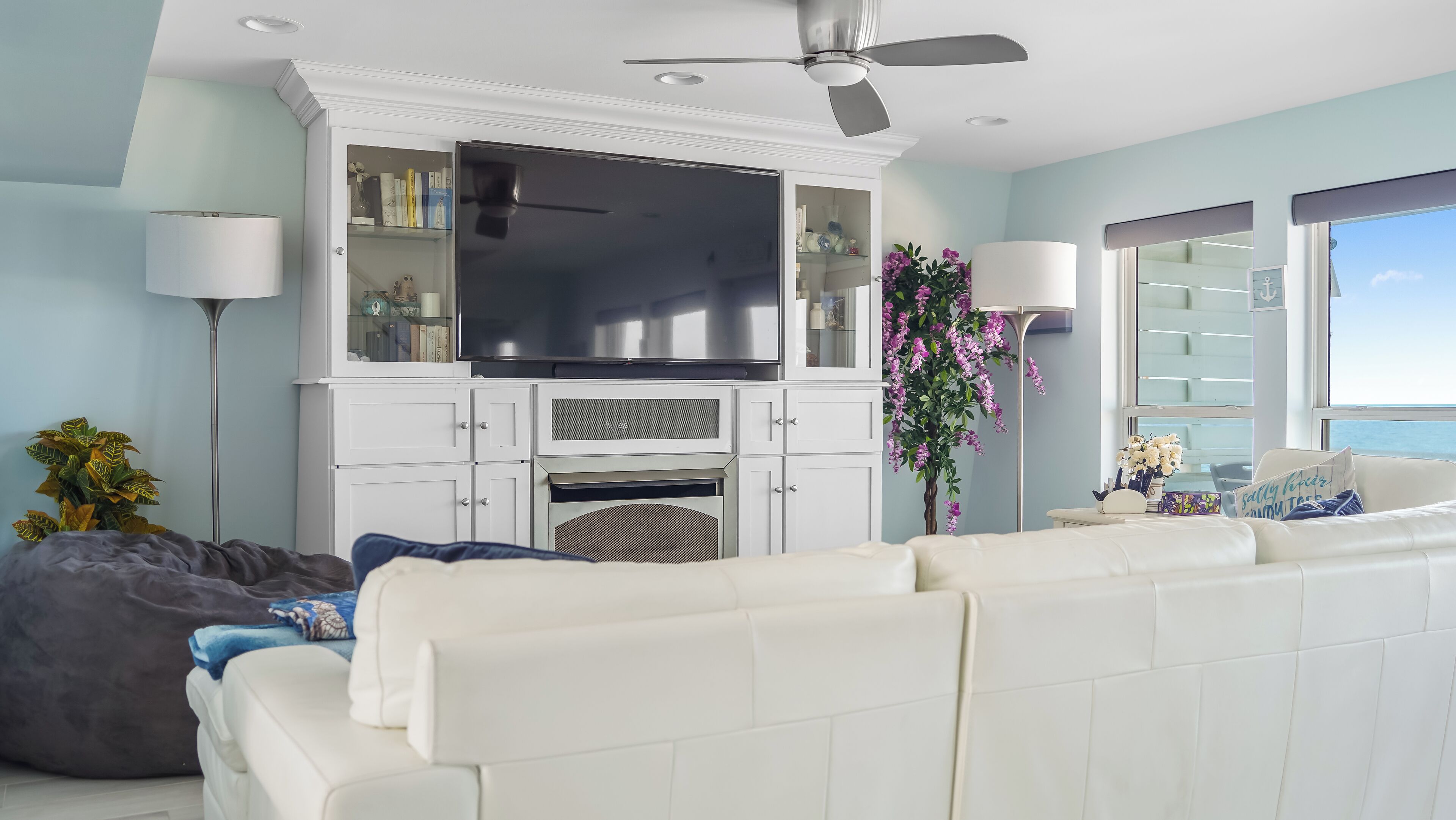 A modern living room with a flat-screen TV, white cabinetry, a ceiling fan, and a large window offering a view of the sea.