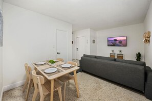 A modern living room with a grey sofa, a dining table set with plates and glasses, and a television mounted on the wall displaying a scenic image.