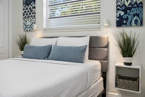 A neatly arranged bedroom with a bed, pillows, a nightstand, a potted plant, and artwork on the walls.