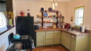 Cocina compacta con refrigerador, estufa y lavamanos. Hay repisas con vajilla y una ventana con cortinas.