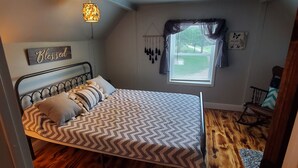 Un dormitorio con cama, ventana con cortinas, suelo de madera y una decoración colgante.