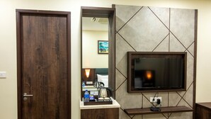 Habitación de hotel con puerta de madera, un espejo, un televisor montado en la pared y un pequeño estante de madera con una tetera y tazas.