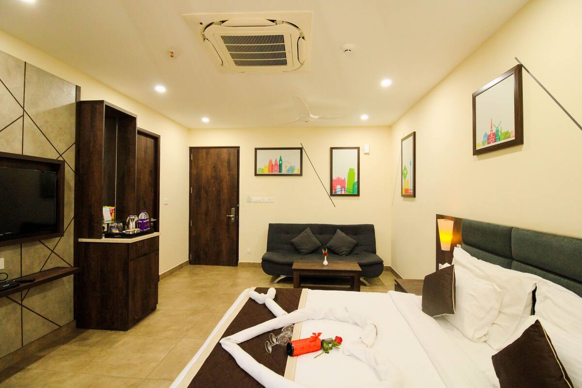 A hotel room with a bed, a TV, a desk, a sofa, and artwork on the walls.
