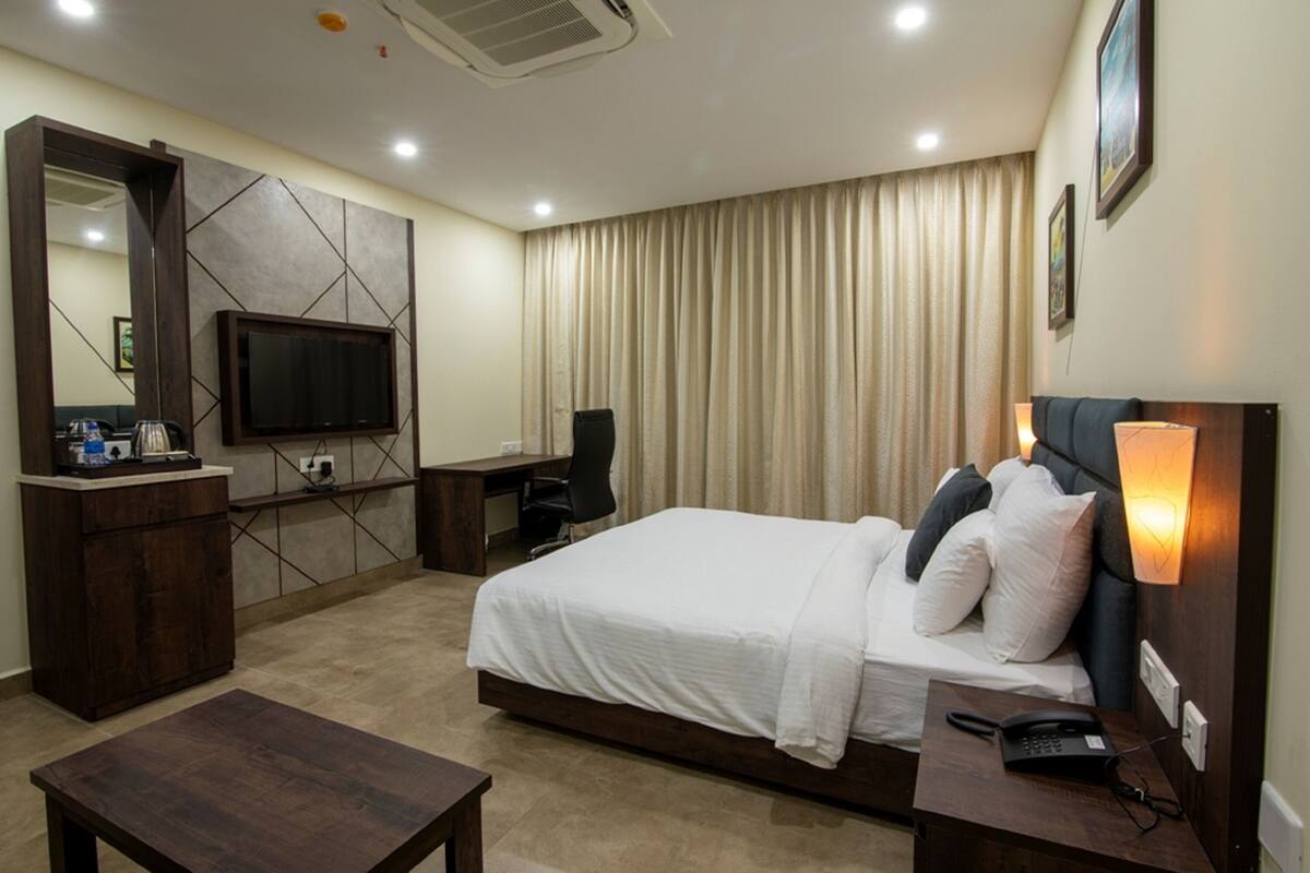 A hotel room with a bed, bedside table, desk, chair, TV, and a large window with curtains.