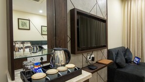 Una habitación de hotel moderna con un televisor de pantalla plana montado en la pared, una zona de café con hervidor y tazas, un sofá con una computadora portátil y un espejo que refleja la decoración de la habitación.